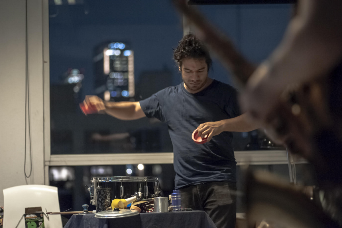 Miguel Manríquez, der vor einem Fenster, hinter dem verschwommen die Lichter eines Hochhauses zu erahnen sind, Musik macht, indem er Klangschalen über einer Trommel bewegt