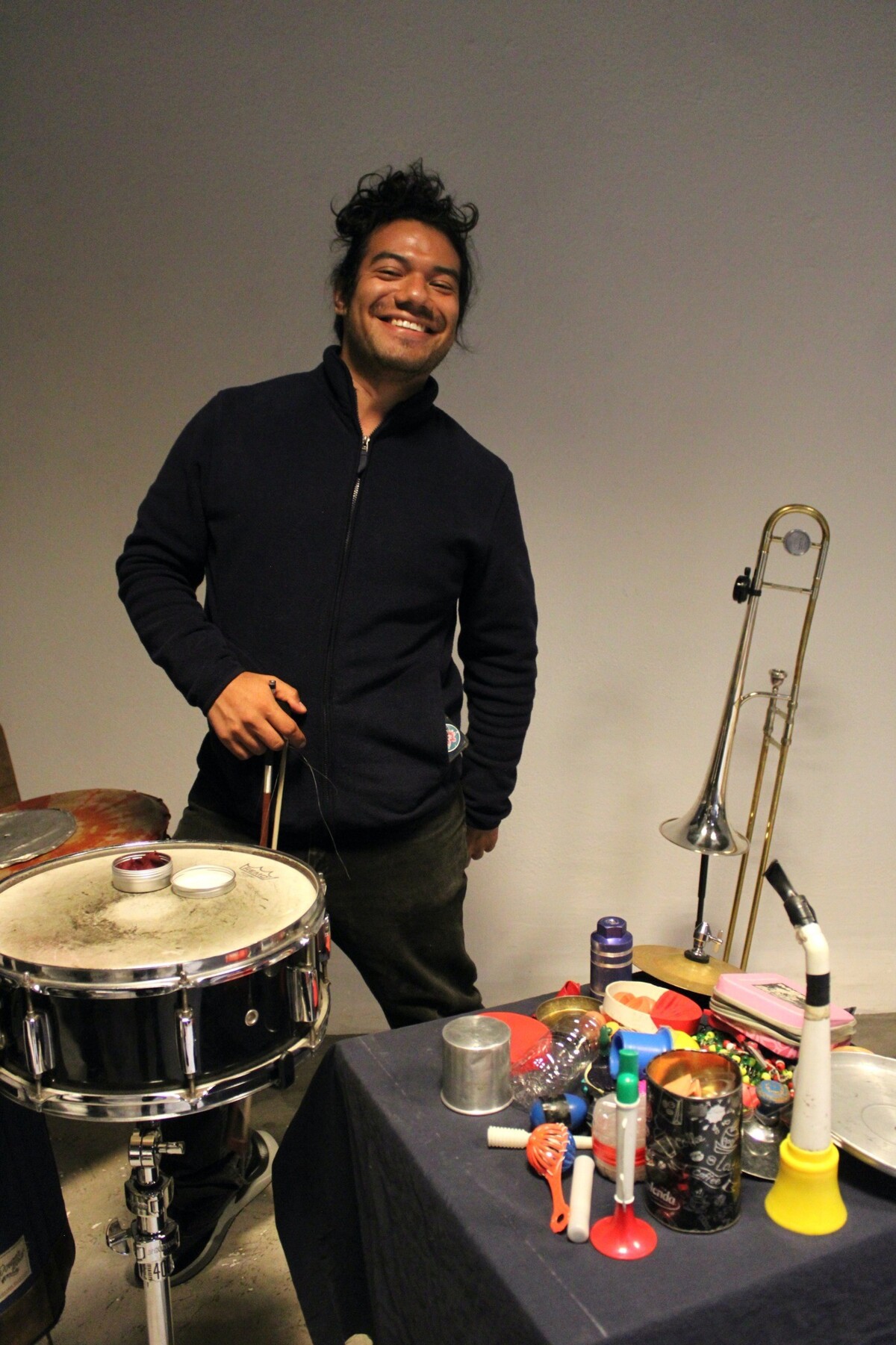 Miguel Manríquez, der vor einer Trommel steht und vielen verschiedenen Alltagsgegenständen, die als Musikinstrumente verwendet werden. Er hält eine Bogen in der Hand und lächelt sympathisch.