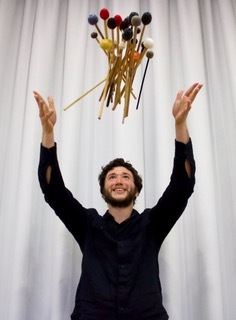 Noah Rosen wirft vor einem weissen Vorhang viele verschiedenen Schlagzeugschläger in die Luft und schaut mit Freude zu dem bunten Wirrwar in der Luft hoch