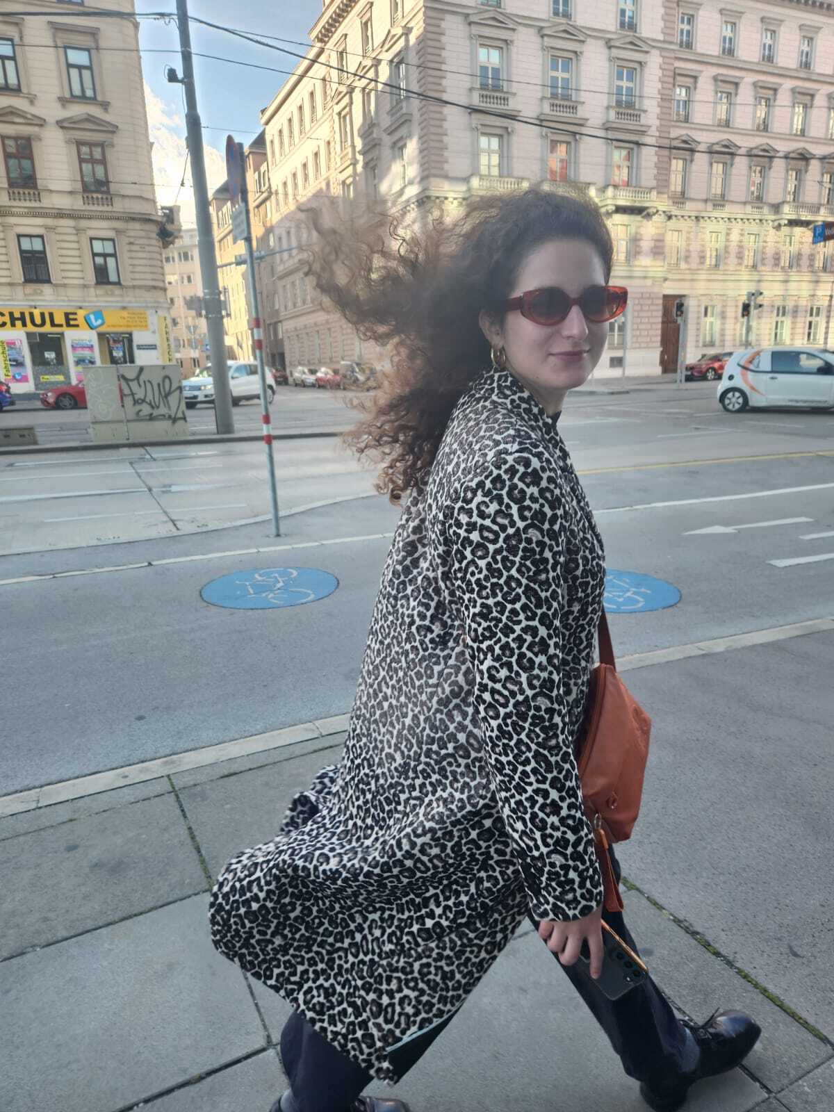 Carmel Curiel läuft durch die Strassen einer Stadt, trägt einen Mantel im Leopardenmuster und eine orange Sonnenbrille