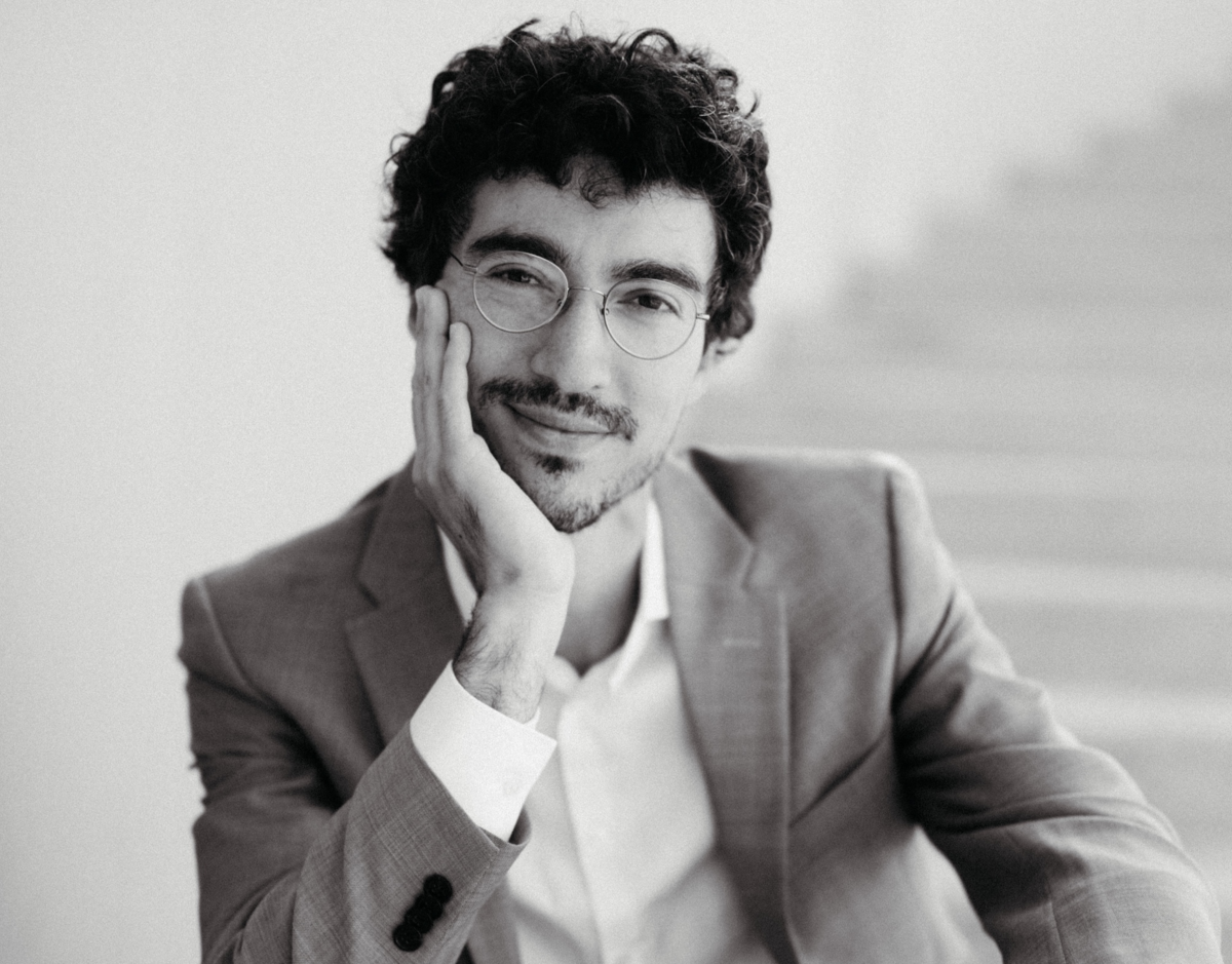 schwarz weiss Foto von Camilo Bornstein, der auf einer Treppe sitzt und sein Gesicht in einer Hand abstützt und in die Kamera lächelt