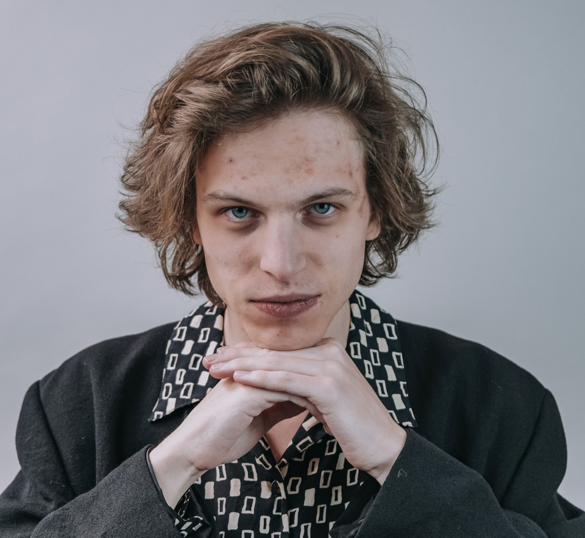 Portrait von Vili Polajnar, der vor grauem Hintergrund in schwarzem Jackett und gemustertem Hemd sein Kinn auf seinen Händen abstützt und frech in die Kamera schaut