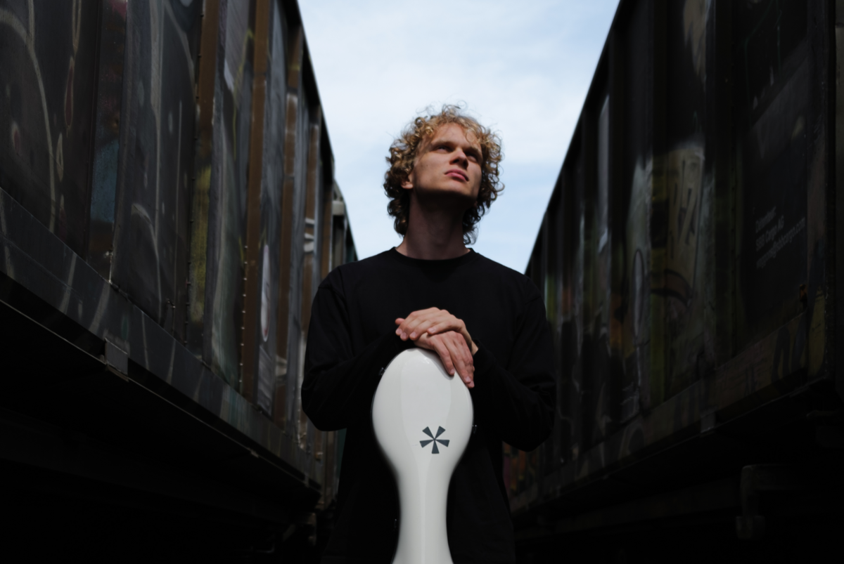 Maksim Barbash steht zwischen zwei dunklen Wänden, über ihm strahlt der blaue Himmel, er stützt seine Hände auf seinem weissen Cello Koffer ab und schaut verträumt nach oben.