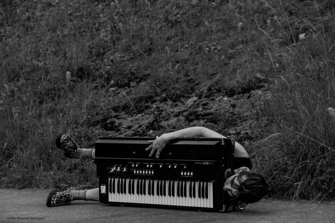 Raphaëlle Proust liegt mit ihrem Keyboard auf dem Boden, dabei hält sie es vor ihrem Körper und leigt seitlich, sodass es aussieht, als ob sie ein vermenschlichtes Keyboard ist.