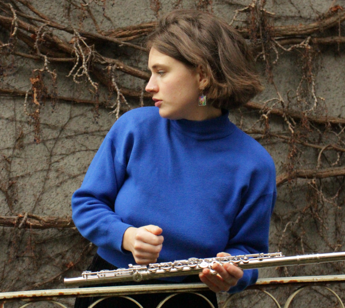 Seraina Ramseier steht vor einer umrankten Betonwand, hält ihre Querflöte in der Hand und schaut rechts aus dem Bild raus.