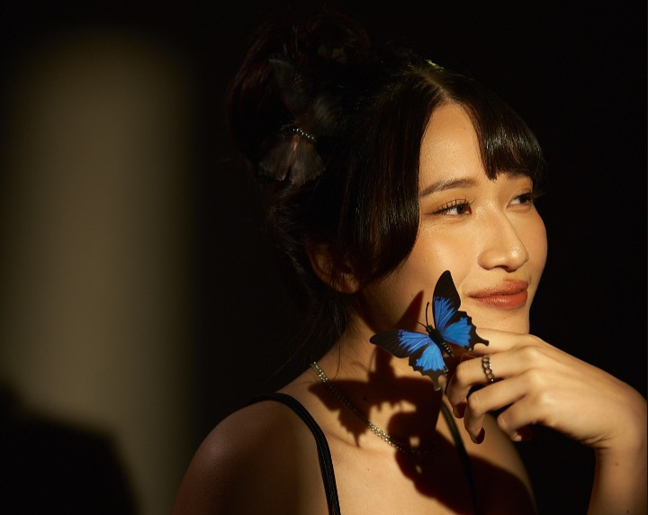 Yu-Yun Peng befindet sich in einem sehr dunklen Raum, während ein Lichtstrahl genau auf sie trifft, sie stützt ihr Kinn in einer Hand, auf dem ein blauer Schmetterling sitzt und schaut aus dem Bild raus.