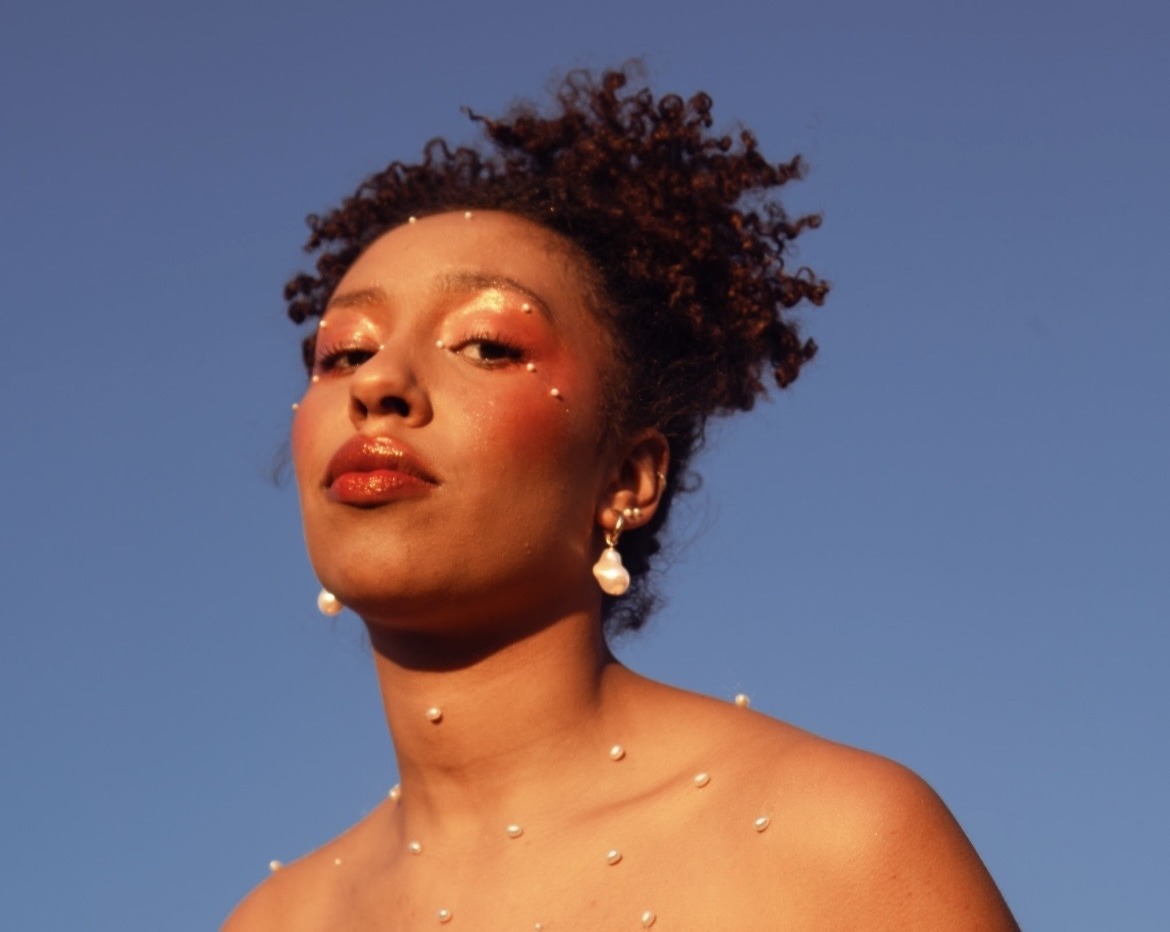 Emily Adomah mit weissen Perlen auf ihrem Körper vor himmelblauem Hintergrund fotographiert aus einer tiefergelegenen Perspektive