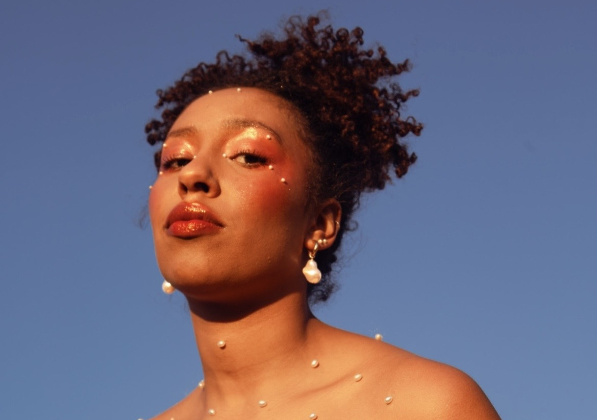 Emily Adomah mit weissen Perlen auf ihrem Körper vor himmelblauem Hintergrund fotographiert aus einer tiefergelegenen Perspektive