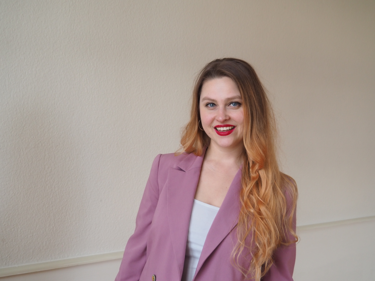 Barbora Jirásková steht vor einem weiss neutralen Hintergrund, von dem sich ihr rosa farbenes Jackett abhebt und lacht sympathisch in die Kamera