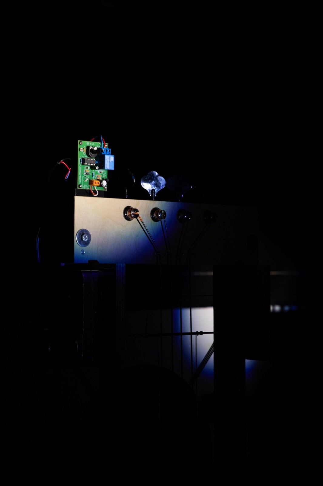 aus dem Schwarz, das das Bild beinahe vollkommen ausmacht, ist eine kleine elektrische grüne Platte zu sehen, die an der Holzplatte unter ihr befestigt ist und mit ihr verbunden ist.