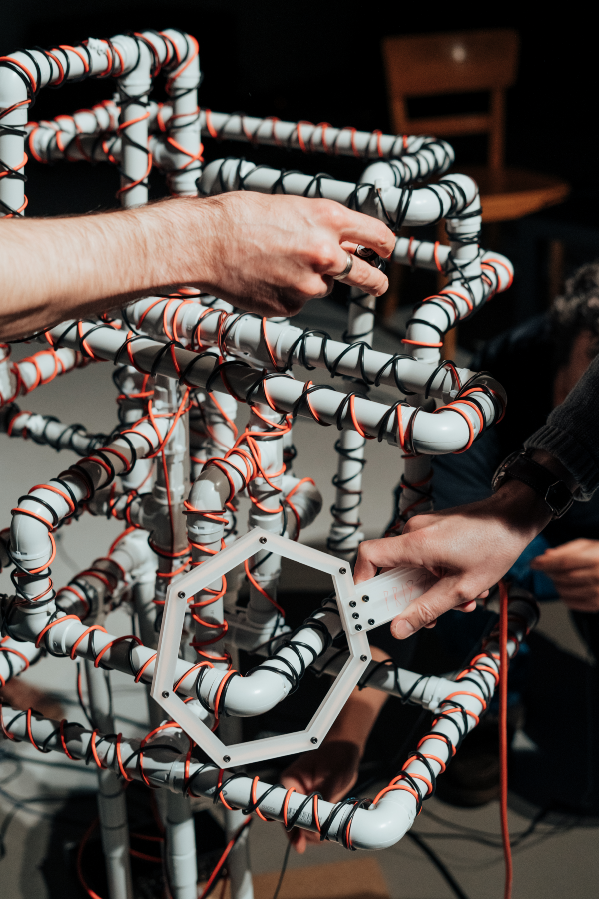 zwei Hände hantieren an der Installation «overengineered», die aus Plastikröhren besteht, die in Kabel eingewickelt sind, welche mit einem Laptop verbunden sind.