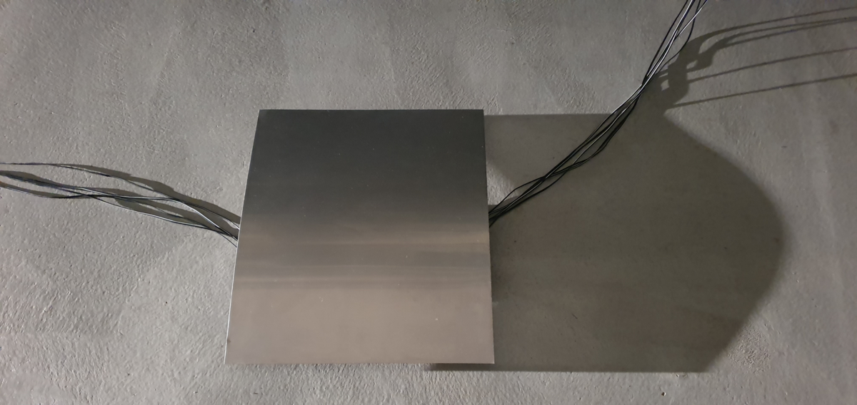 Das Bild zeigt eine der Metallplatten genauer, sie liegt auf einem grauen Boden, ist leicht gewölbt und links und rechts kommen Kabel unter der Wölbung hervor.