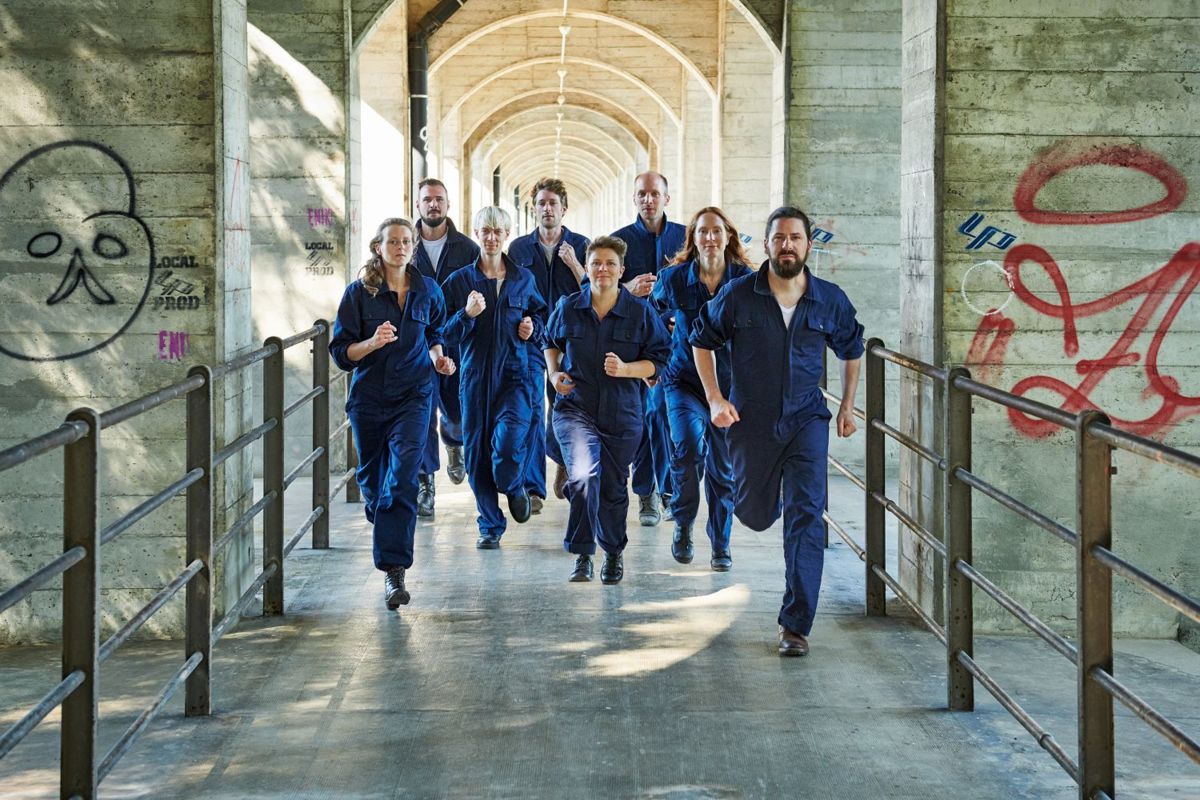 Foto: Zu sehen sind die Mitglieder des Ensemble Proton Bern in blauen Overalls. Sie rennen alle auf die Kamera zu.