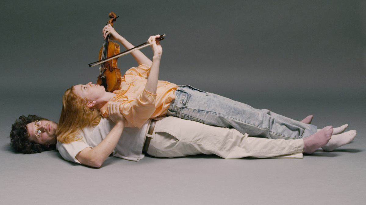 Foto: Zu sehen sind Noëlle-Anne Darbellay und Samuel Stoll. Beide liegen auf dem Boden. Noëlle-Anne Darbellay scheint im Liegen Geige zu spielen.