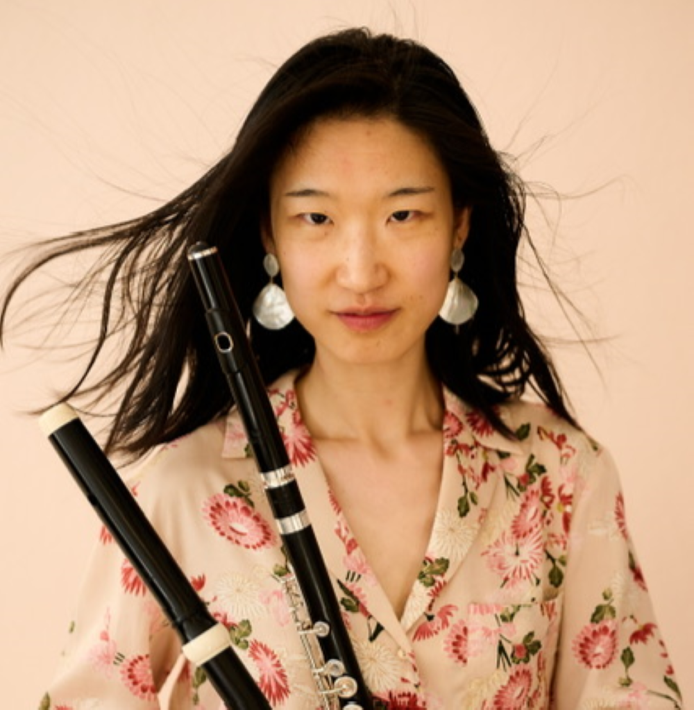 Juli (Xueqing) Wang steht vor einem hellrosa Hintergrund mit wehenden Haaren und hält ihre zwei Flöten in der Hand